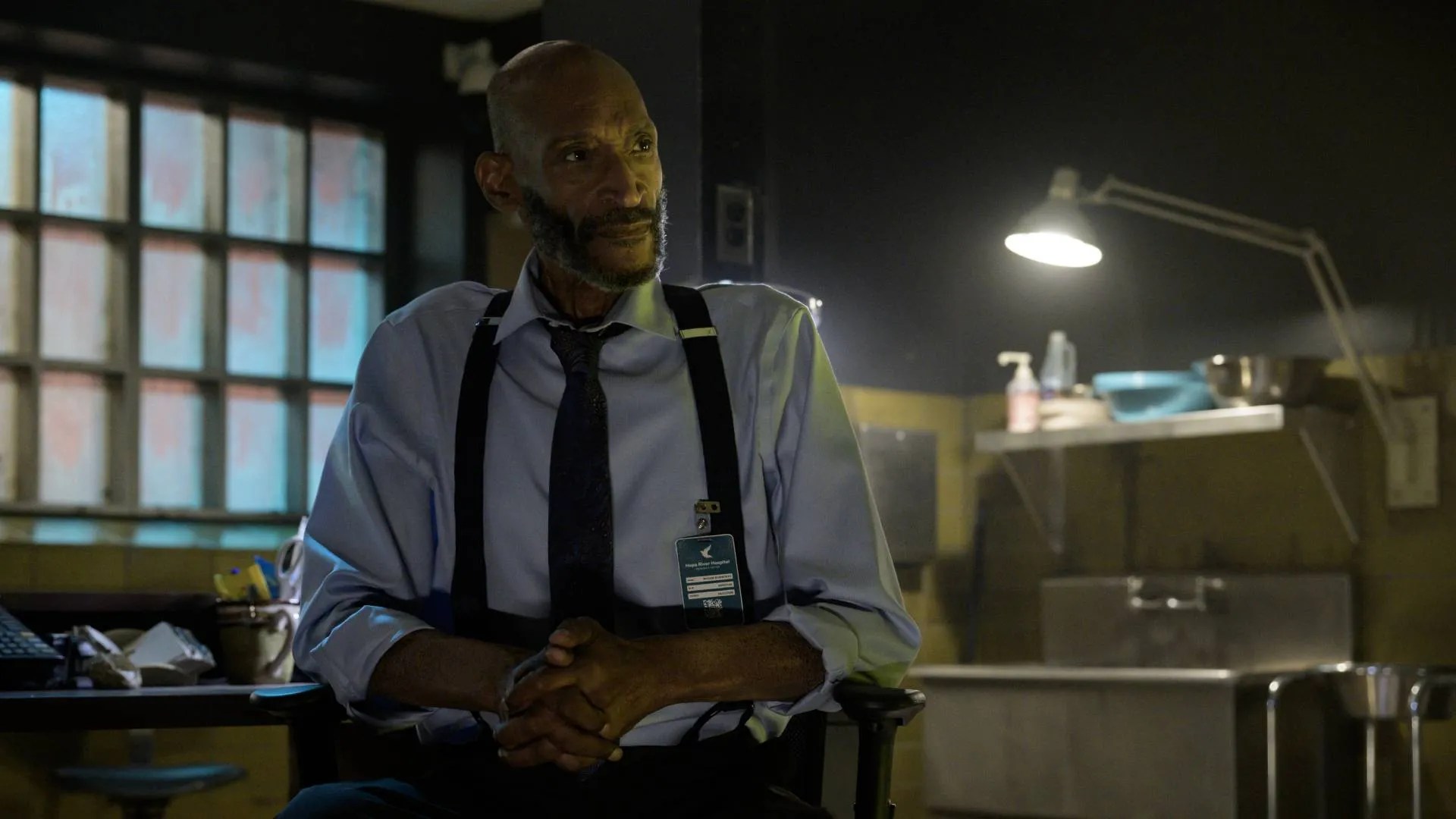Tony Todd sits in a dimly lit office, wearing a light blue dress shirt, dark tie, and suspenders, with an official badge clipped to his pocket.