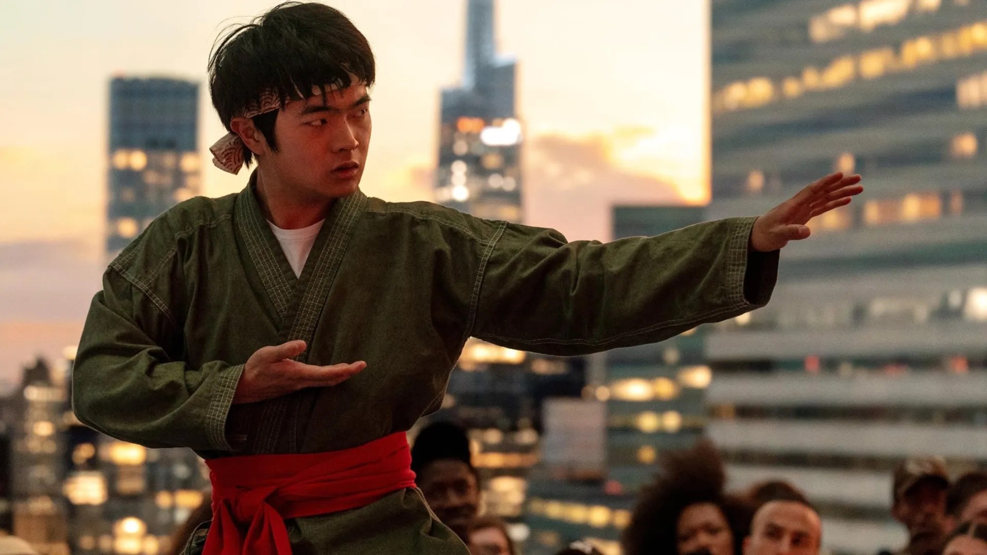 Ben Wang stands in a karate pose wearing a traditional martial arts uniform with a red belt, set against a glowing sun and cityscape silhouette in the promotional poster for Karate Kid Legends.