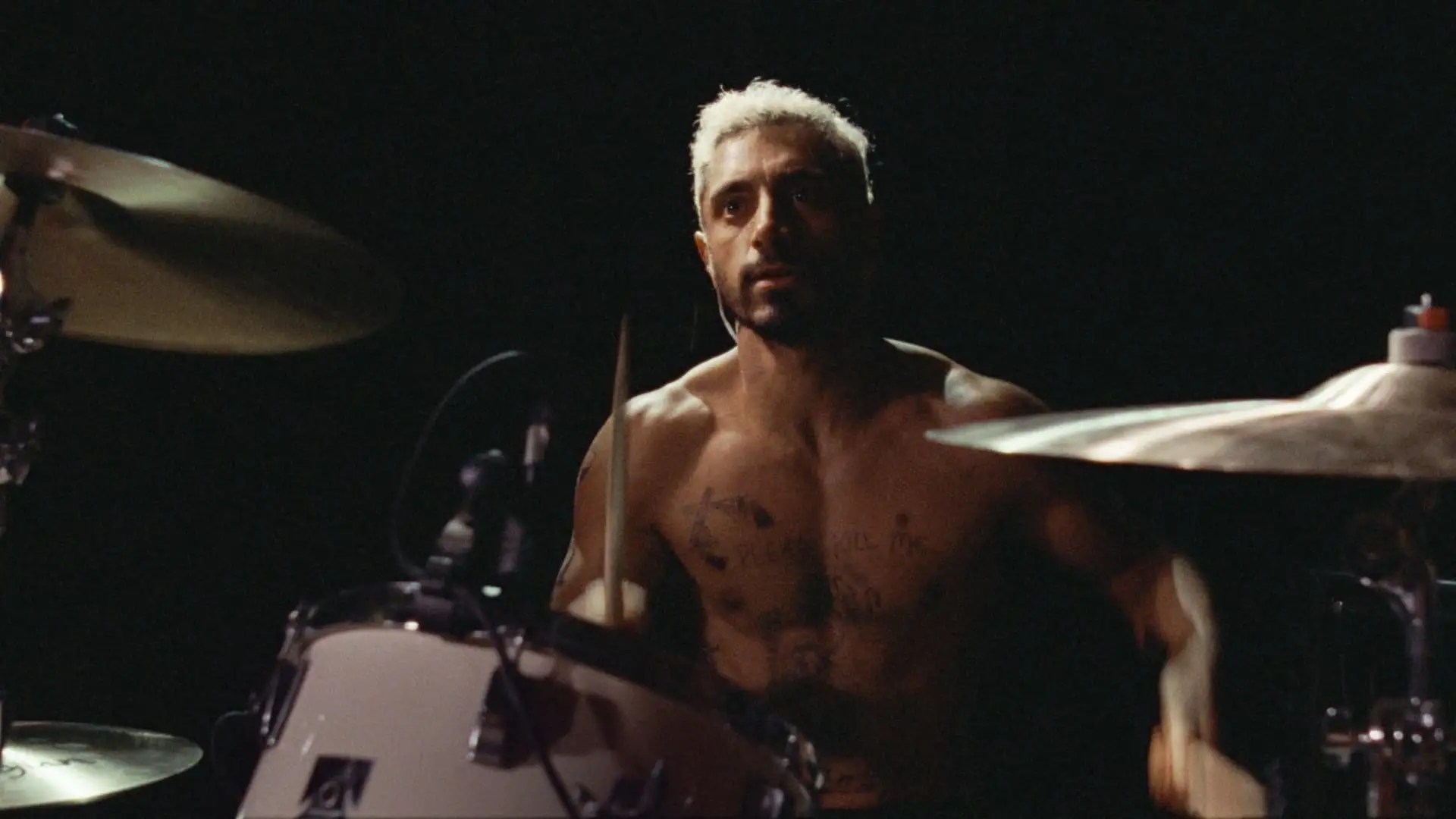 Ruben Stone, played by Riz Ahmed, plays the drums intensely, shirtless with tattoos, illuminated in a dark setting.