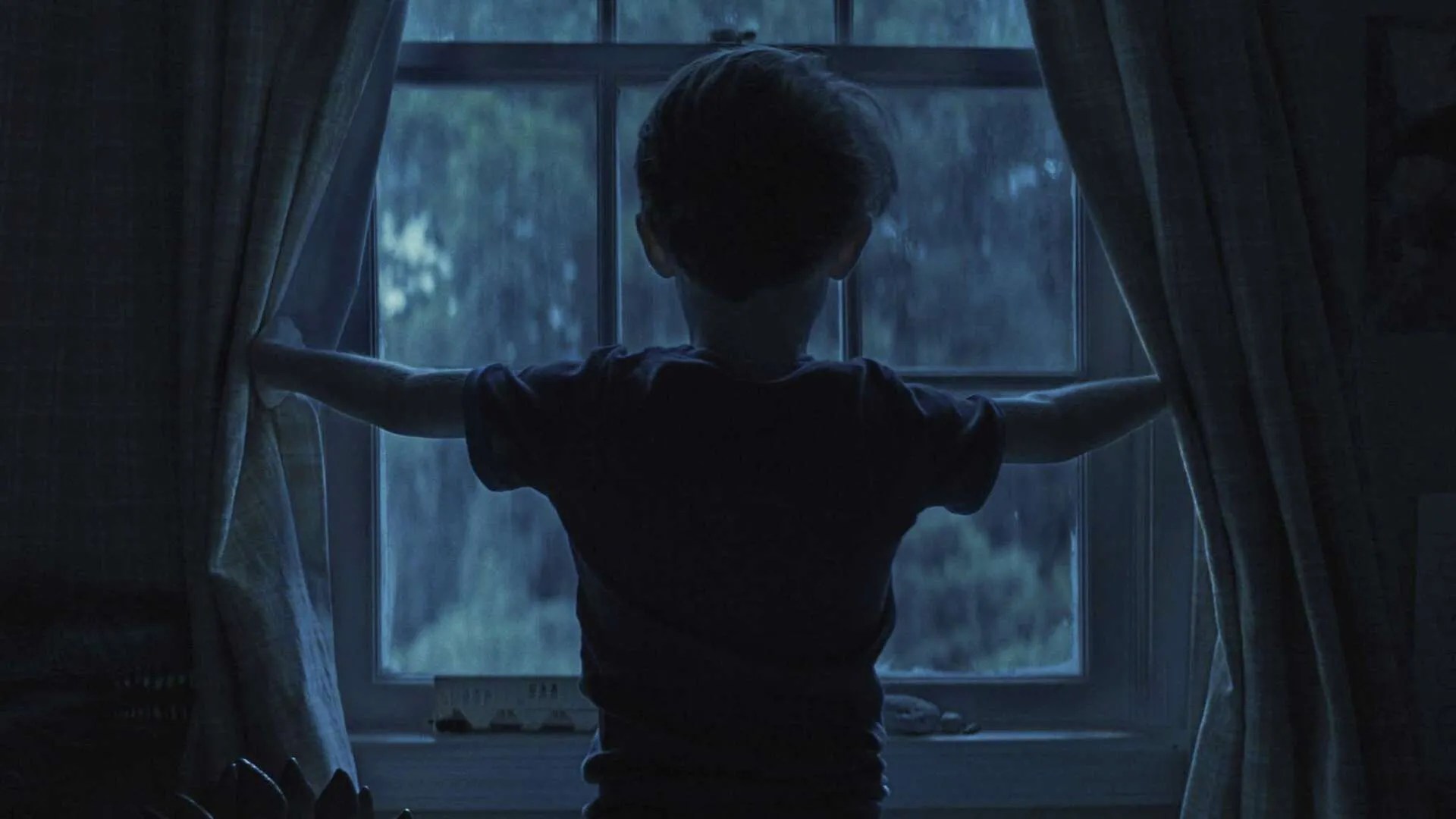 Weapons (2025) Movie Screenshot - Alex (Cary Christopher) stood at a window at night, holding the curtains open.
